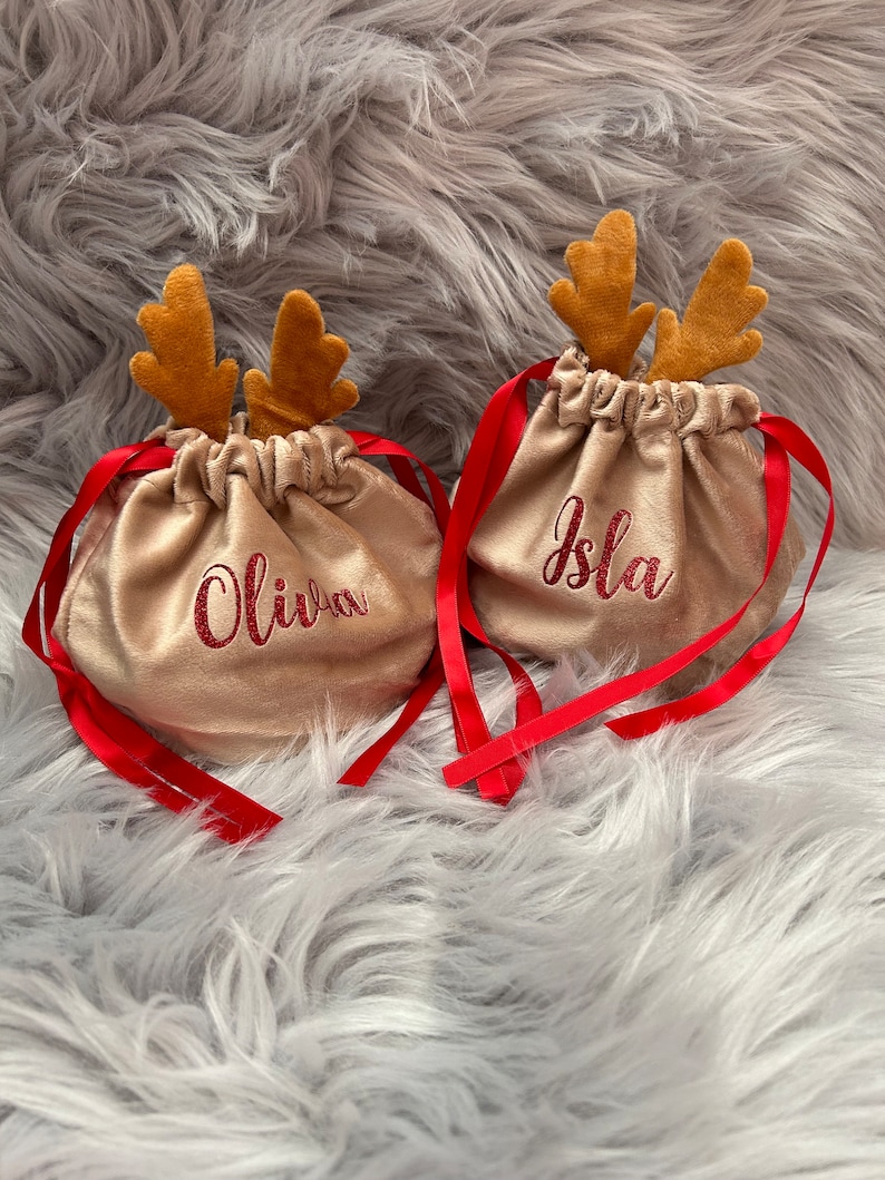 May include: Two gold-colored drawstring bags with red ribbon ties and brown reindeer antlers. The bags have the names "Olivia" and "Isla" embroidered in red glitter. The bags are on a fluffy, gray surface.