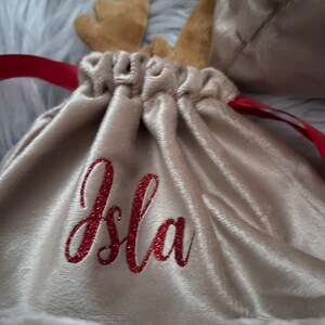 May include: A beige velvet drawstring bag with red glitter text that reads "Isla". The bag has brown felt reindeer antlers and a red ribbon drawstring. A second bag is visible in the background.