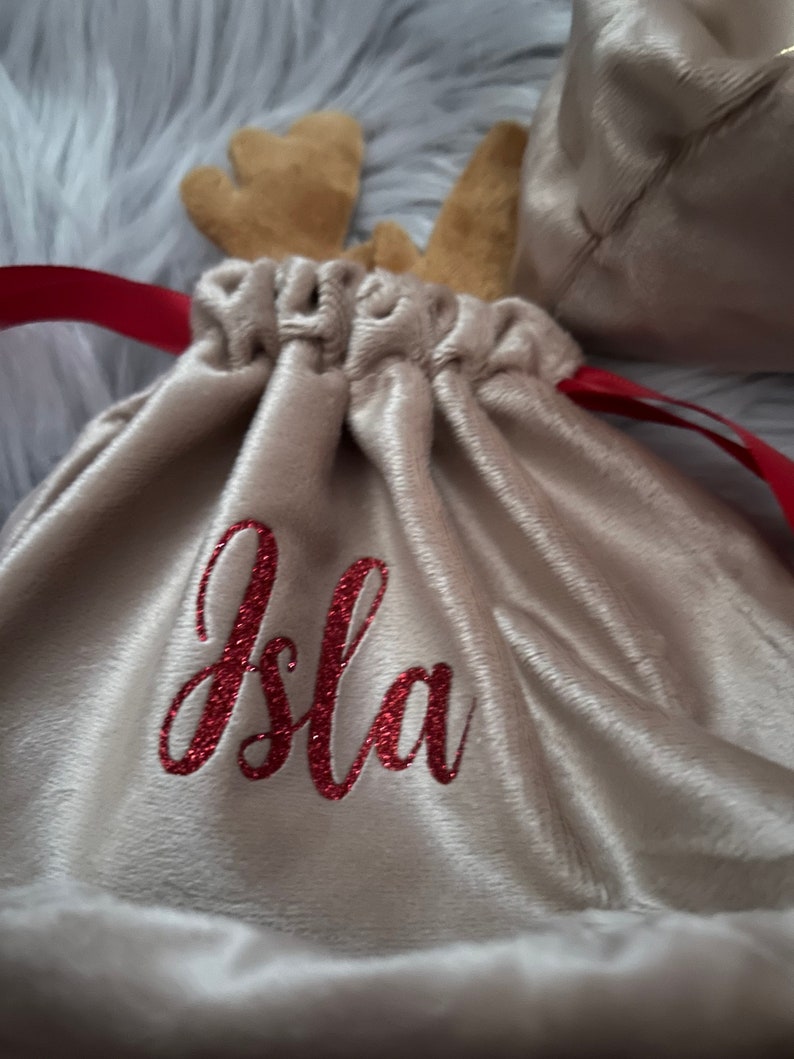 May include: A beige velvet drawstring bag with red glitter text that reads "Isla". The bag has brown felt reindeer antlers and a red ribbon drawstring. A second bag is visible in the background.