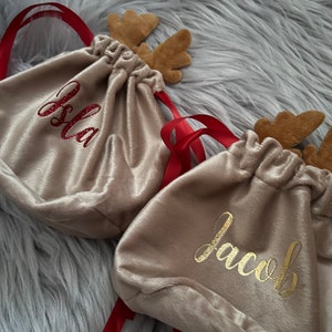 May include: Tan velvet drawstring bags with red ribbon ties and felt reindeer antlers. One bag has the name "Isla" in red glitter, and another has "Jacob" in gold script. The bags are on a gray, fuzzy surface.