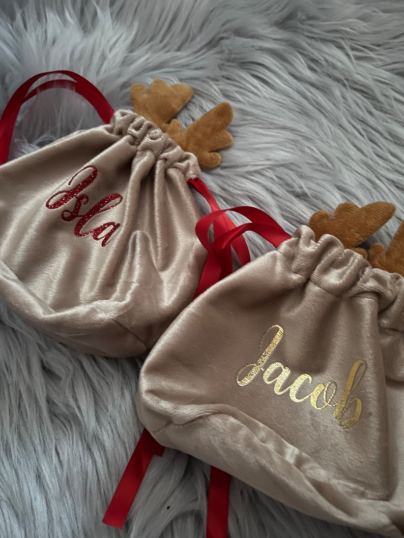 May include: Tan velvet drawstring bags with red ribbon ties and felt reindeer antlers. One bag has the name "Isla" in red glitter, and another has "Jacob" in gold script. The bags are on a gray, fuzzy surface.