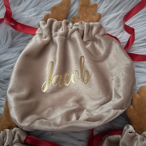 May include: Tan velvet drawstring gift bags with reindeer antlers and red ribbon. The word "Jacob" is printed in gold script on the front of the bag. The bags are on a fluffy, gray surface.
