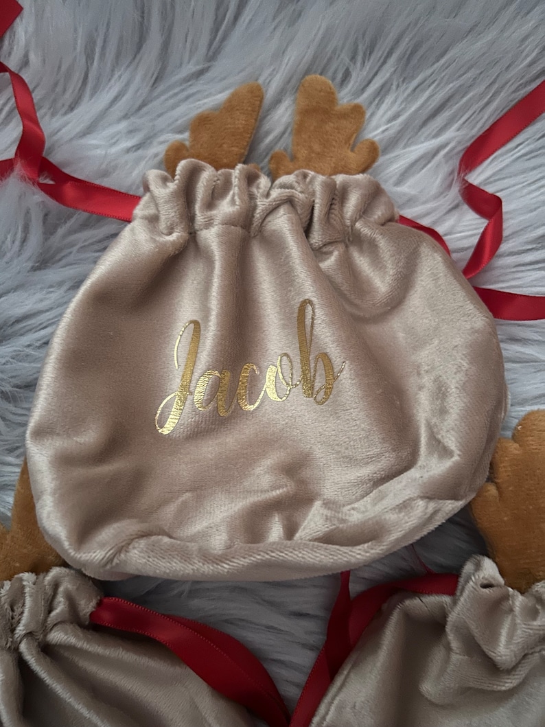 May include: Tan velvet drawstring gift bags with reindeer antlers and red ribbon. The word "Jacob" is printed in gold script on the front of the bag. The bags are on a fluffy, gray surface.