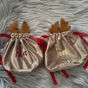 May include: Two beige velvet drawstring bags with brown reindeer antlers and red ribbons. One bag has the name "Isla" embroidered in red, and the other has "Jacob" in gold. The bags are on a fluffy gray surface.