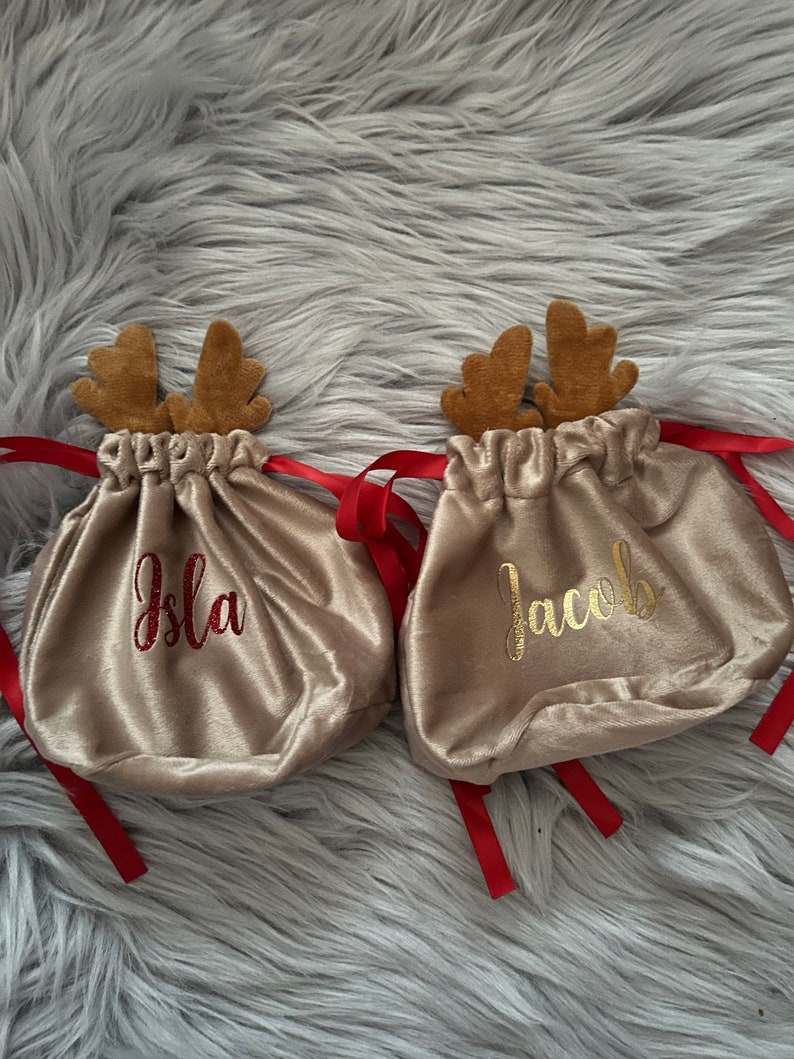 May include: Two beige velvet drawstring bags with brown reindeer antlers and red ribbons. One bag has the name "Isla" embroidered in red, and the other has "Jacob" in gold. The bags are on a fluffy gray surface.