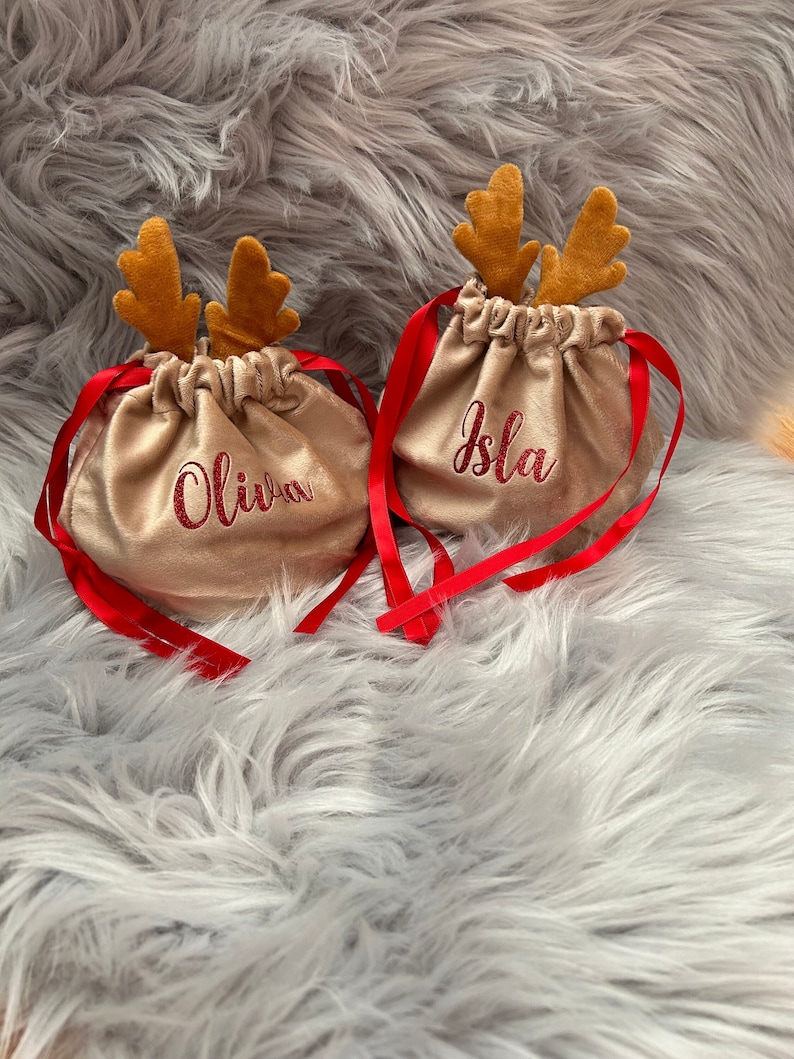 May include: Two beige drawstring gift bags with red ribbon ties and embroidered names, "Olivia" and "Isla". Each bag has brown reindeer antlers attached. The bags are on a fluffy, gray surface.