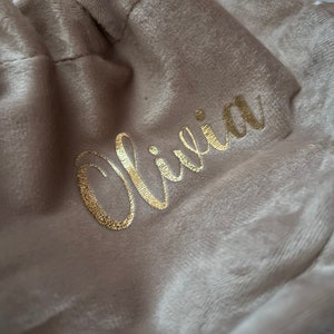 May include: A beige velvet drawstring bag with the name "Olivia" in gold script. The bag is gathered at the top, and the soft fabric has a subtle sheen. The background is a blurred, light-colored surface.