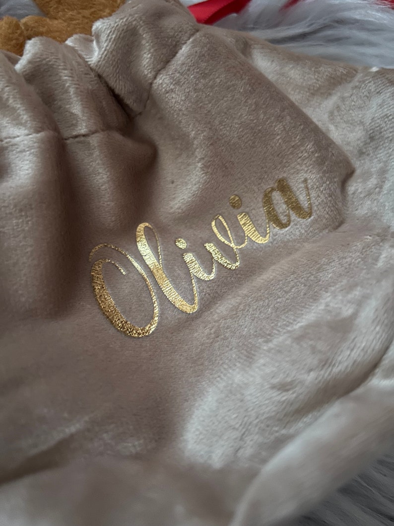 May include: A beige velvet drawstring bag with the name "Olivia" in gold script. The bag is gathered at the top, and the soft fabric has a subtle sheen. The background is a blurred, light-colored surface.