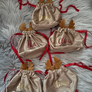 May include: Five beige velvet drawstring bags with red ribbon ties and attached reindeer antlers. Each bag has a name embroidered in gold or red: Jacob, Isla, Olivia, Elliott, and Olivia. The bags are on a fluffy gray surface.