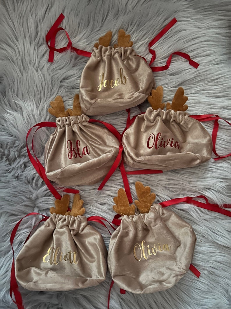 May include: Five beige velvet drawstring bags with red ribbon ties and attached reindeer antlers. Each bag has a name embroidered in gold or red: Jacob, Isla, Olivia, Elliott, and Olivia. The bags are on a fluffy gray surface.