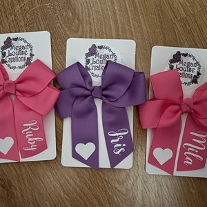 May include: Three hair bows with personalised names, "Ruby", "Iris", and "Mila", on white cards with a logo for "Megan Louise Creations".