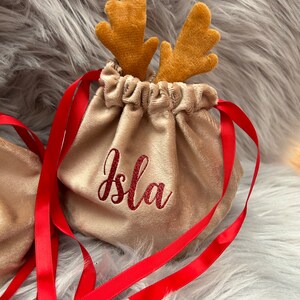 May include: A beige velvet drawstring bag with red ribbon ties and embroidered name "Isla". The bag is topped with plush reindeer antlers. Festive and decorative.