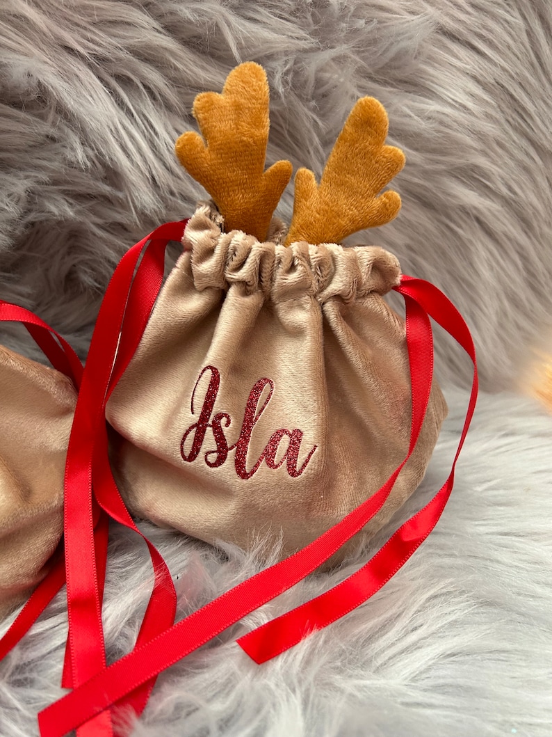 May include: A beige velvet drawstring bag with red ribbon ties and embroidered name "Isla". The bag is topped with plush reindeer antlers. Festive and decorative.