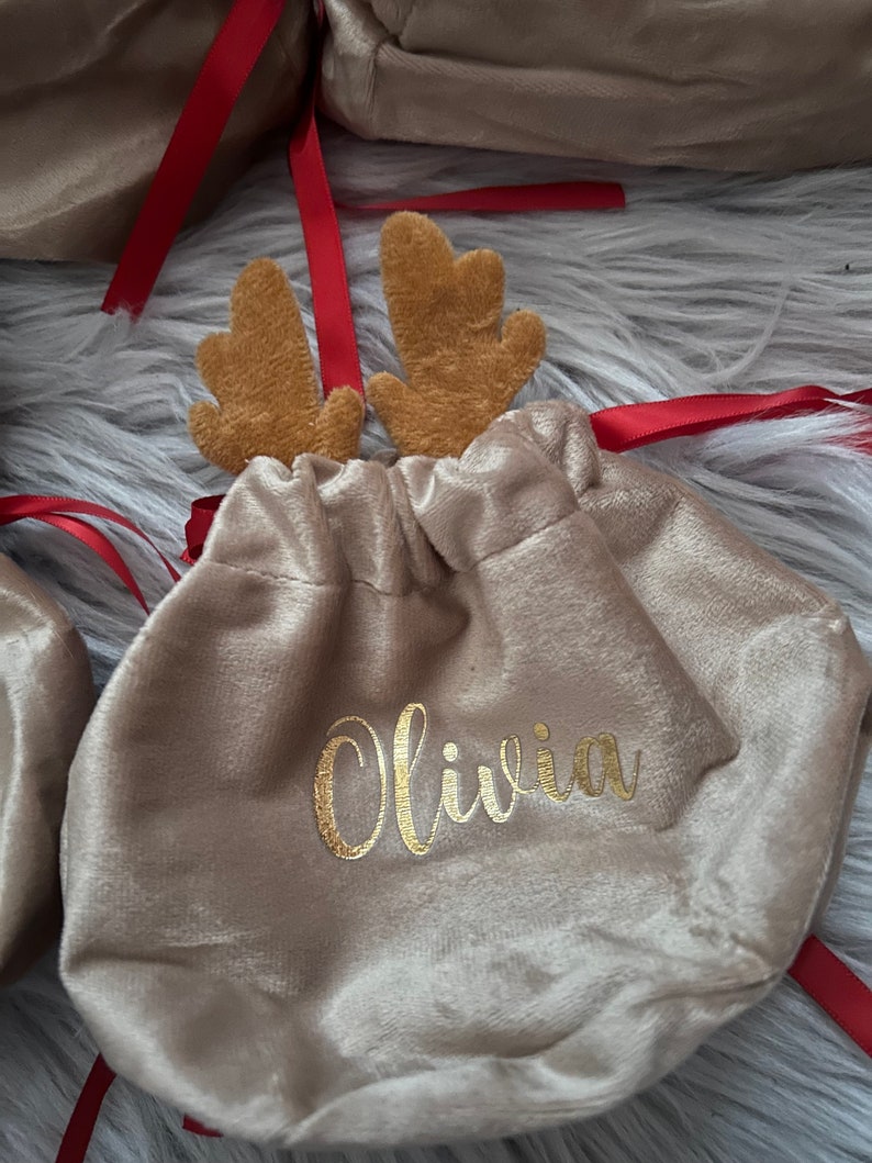 May include: Tan velvet gift bag with gold lettering that reads "Olivia". The bag is adorned with brown reindeer antlers and red ribbon. The bag is closed with a drawstring.