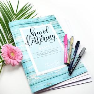 Może przedstawiać: Turkusowa książka z wzorem drewna i tekstem "hand lettering basics" w czarnym stylu pisma odręcznego. Książka nosi tytuł "Anleitungs- und &Uuml;bungshet f&uuml;r Brushpen und Fineliner" i zawiera imię "Lutorila Biem" w stylu pisma odręcznego. Książka leży na białym tle z r&oacute;żowym kwiatem i kilkoma czarnymi i szarymi długopisami.