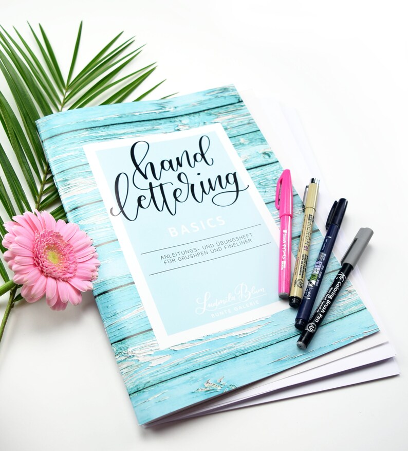 Może przedstawiać: Turkusowa książka z wzorem drewna i tekstem "hand lettering basics" w czarnym stylu pisma odręcznego. Książka nosi tytuł "Anleitungs- und &Uuml;bungshet f&uuml;r Brushpen und Fineliner" i zawiera imię "Lutorila Biem" w stylu pisma odręcznego. Książka leży na białym tle z r&oacute;żowym kwiatem i kilkoma czarnymi i szarymi długopisami.