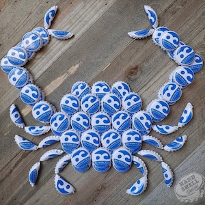 May include: A blue and white crab made from bottle caps. The crab is made from bottle caps from a brewery called "Brewing Co.". The crab is on a wooden background.