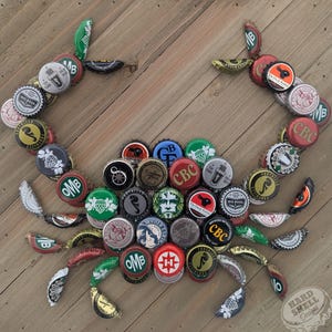 May include: A crab made from bottle caps, with a variety of colors and designs. The crab is on a wooden background. The text "HARD SHELL Designs" is in the lower right corner.