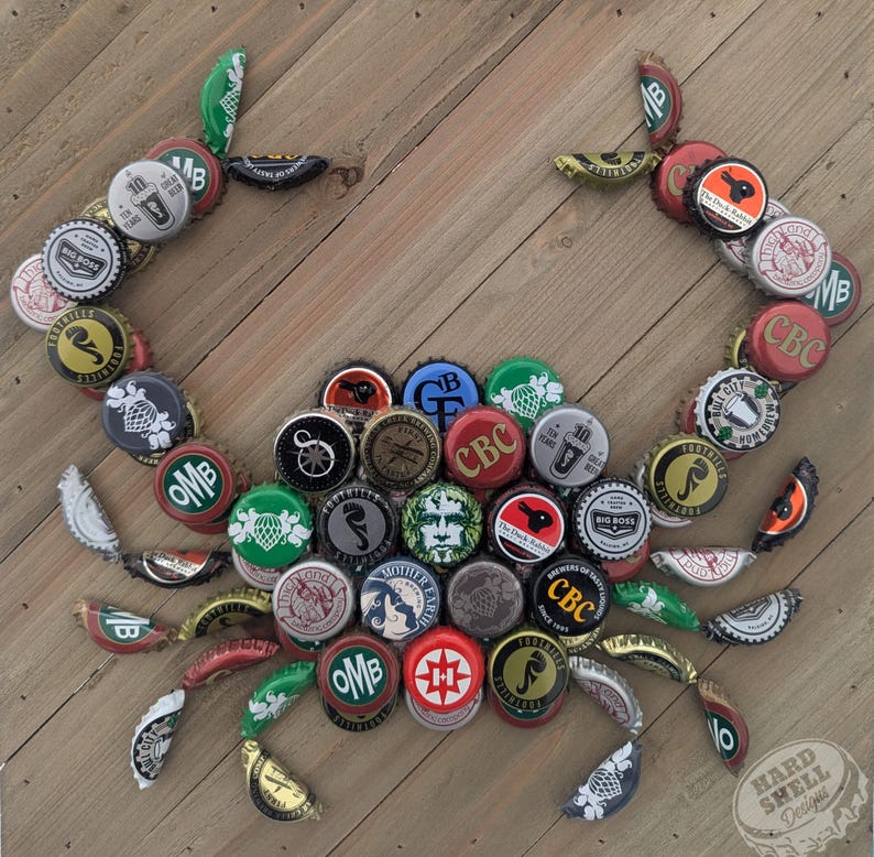 May include: A crab made from bottle caps, with a variety of colors and designs. The crab is on a wooden background. The text "HARD SHELL Designs" is in the lower right corner.