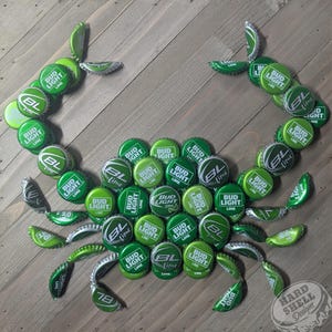Puede incluir: Un cangrejo verde hecho con tapas de botellas de Bud Light Lime sobre un fondo de madera. El cangrejo está hecho con las tapas de botellas que forman el cuerpo y las patas. El cangrejo está mirando hacia la derecha. Las palabras "Hard Shell Designs" están en la esquina inferior derecha de la imagen.