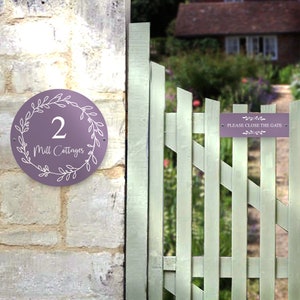 May include: Purple round house number sign with white floral design and the number 2 and the text "Mill Cottages". A white sign with the text "Please close the gate" is attached to a white picket fence.