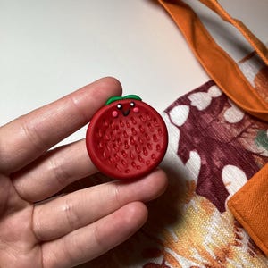 Puede incluir: Un dije rojo con forma de fresa con una cara sonriente y hojas verdes. El dije tiene una superficie texturizada y se sostiene en una mano. El fondo incluye un bolso tote naranja y una tela con estampado floral.