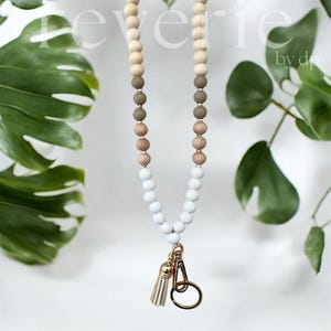 May include: A wooden bead lanyard with a tassel and gold-coloured hardware. The lanyard has a gradient of wooden beads, changing from natural wood tones to grey and white. The word "reverie" is visible at the top.