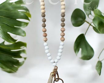 Puede incluir: Un cordón con cuentas con una borla y herrajes dorados. El cordón presenta un degradado de cuentas de madera, que transiciona de tonos de madera natural a gris y blanco. La palabra "reverie" es visible en la parte superior.