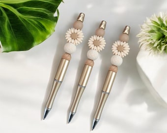 May include: Three ballpoint pens with metallic gold barrels and silver tips. Each pen features a decorative topper consisting of a smooth wooden bead, a textured white bead, a beige flower-shaped bead, and a smooth beige bead with a gold cap.
