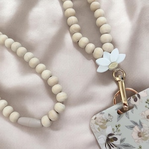 May include: A lanyard featuring light-coloured wooden beads strung together, accented by a white silicone flower charm. A gold-toned metal lobster clasp connects to a rectangular badge holder decorated with a floral pattern in shades of green and white.