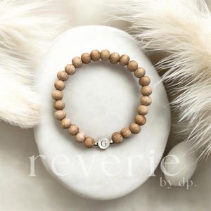 Könnte beinhalten: Ein Armband aus Holzperlen mit einem silbernen Charm mit dem Buchstaben "G". Das Armband wird auf einer weißen, oval geformten Oberfläche präsentiert, mit weichen, federartigen Elementen im Hintergrund. Das Wort "reverie" ist auf der Oberfläche aufgedruckt.