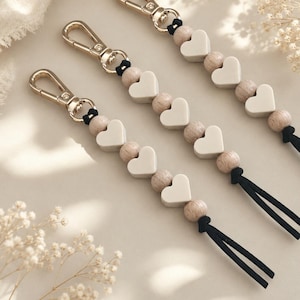 May include: Three keychains featuring gold-toned metal lobster clasps, alternating light beige heart-shaped beads and natural wooden spherical beads, strung on black cord. Each keychain ends with a black cord tassel at the bottom.