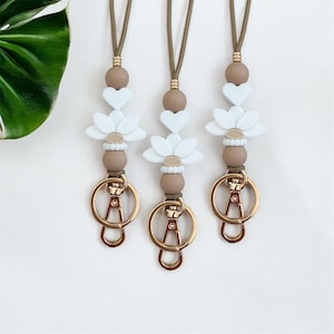 Peut inclure: Trois lanières avec un cordon marron clair, chacune avec un fermoir doré. Les lanières ont un motif de cœurs bleu clair, de perles blanches en forme de fleur et de perles marron. Le fond est blanc.