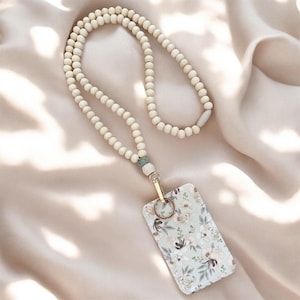 Puede incluir: Un cordón para credencial compuesto por una larga hilera de cuentas de madera de color claro con una cuenta gris como acento. Un broche de metal dorado une el cordón a un porta credencial rectangular con un estampado floral en tonos pastel.