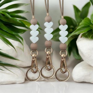 Peut inclure: Trois lanières avec des cordons marron clair, des perles beiges et des perles en forme de cœur blanches. Chaque lanière a un fermoir et un anneau de couleur or. Le fond présente un feuillage vert et des pierres blanches.