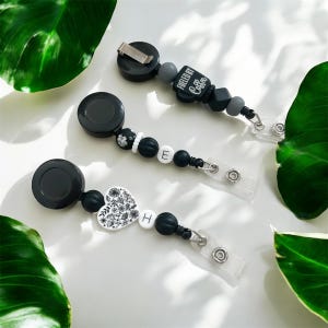 Peut inclure: Trois porte-badges à perles noires et blanches. L'un comporte un charm en forme de cœur avec des détails floraux, un autre la lettre "E", et le troisième le texte "Fueled by Coffee". Chaque enrouleur a un cordon rétractable et un porte-badge en plastique transparent.