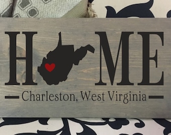 May include: A gray wooden sign with the word "HOME" in black letters. The state of West Virginia is depicted in black with a red heart in the center. The text "Charleston, West Virginia" is written below the state.