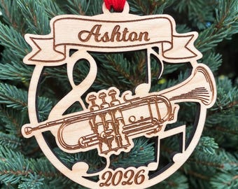 May include: A circular laser-cut wooden ornament featuring a trumpet and musical note design. The top banner is engraved with the name Ashton, and the bottom section displays the year 2026. A red ribbon is attached to the top for hanging.