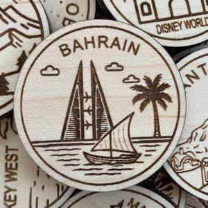May include: A round wooden Bahrain souvenir featuring etched illustrations of the Bahrain World Trade Center, a sailboat, a palm tree, and the word "BAHRAIN". The design is surrounded by a circular border and two clouds.