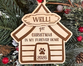 May include: A light-colored wooden dog house ornament with a red ribbon hanger. The text reads WELLI, 1ST CHRISTMAS IN MY FUREVER HOME, and 2026. A paw print is engraved above the year. The ornament is shaped like a house with a sign on top.