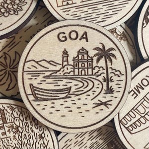 May include: Wooden round coaster with the word "GOA" at the top. The coaster features an etched design of a coastal scene with a church, palm tree, boat, and mountains. The coaster is made of light-colored wood.