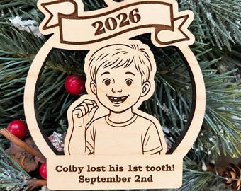 May include: A light-colored wooden ornament featuring a laser-engraved illustration of a child holding a tooth. The top banner displays the year 2026, and the bottom section includes the text: Colby lost his 1st tooth! September 2nd.