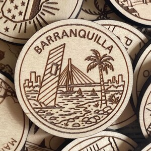 May include: Wooden Barranquilla souvenir featuring a laser-etched design of a bridge, palm tree, and city skyline. The word "Barranquilla" is etched around the edge of the wooden circle. Other wooden circles with etched designs are visible.