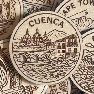 May include: Wooden round featuring a detailed engraving of Cuenca, Spain. The design includes a bridge, buildings, and a mountain range, with the city name arched above. The image is in a natural wood tone.