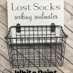 May include: A white wooden sign with black text that reads "Lost Socks seeking soulmates". A black wire basket is attached to the sign. The sign is sitting on a wood surface. The text "White Paint" is written in black letters on the wood surface.