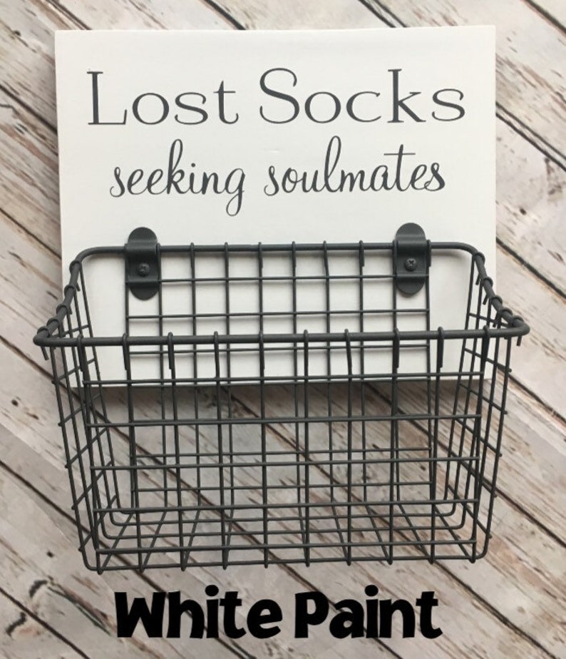 May include: A white wooden sign with black text that reads "Lost Socks seeking soulmates". A black wire basket is attached to the sign. The sign is sitting on a wood surface. The text "White Paint" is written in black letters on the wood surface.