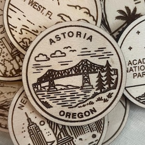 May include: A collection of round wooden coasters with laser-etched designs. One coaster features a bridge, trees, and the text "Astoria Oregon". Other coasters show various landmarks and locations.
