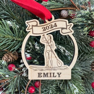 May include: A wooden Christmas ornament featuring a silhouette of a person paddleboarding with the year 2024 and the name "EMILY" engraved on it.