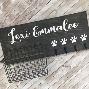 May include: A dark gray wooden sign with the names "Lexi Emmalee" in white cursive lettering. The sign has four white paw prints and five hooks for hanging leashes. A black metal wire basket is attached to the left side of the sign.