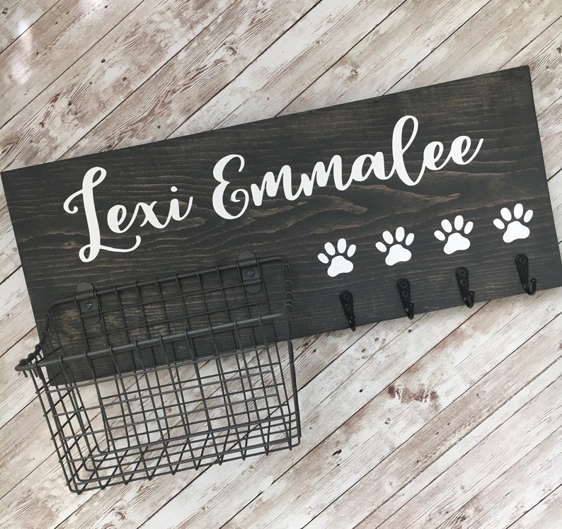 May include: A dark gray wooden sign with the names "Lexi Emmalee" in white cursive lettering. The sign has four white paw prints and five hooks for hanging leashes. A black metal wire basket is attached to the left side of the sign.